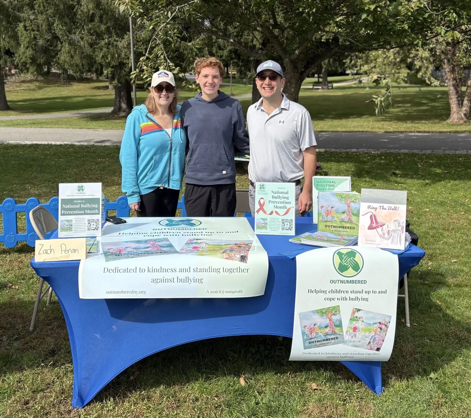 Zachary Penan standing behind the Outnumbered table between two adults. Banners on the table read “Helping children stand up to and cope with bullying” and “Dedicated to kindness and standing together against bullying.”