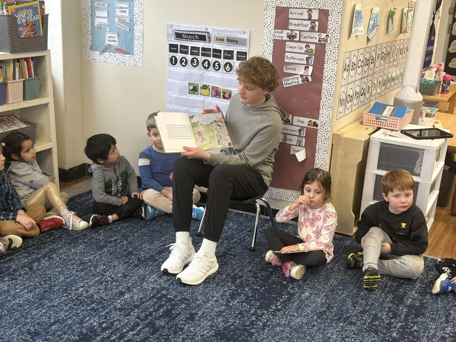 Zachary Penan seated on a classroom chair while reading Outnumbered aloud to pre-k students gathered on a rug at Little Friends in Greenwich.